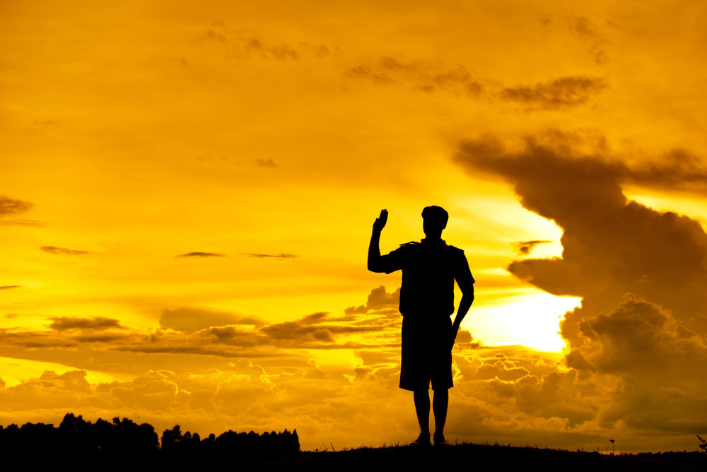 A scout holds up the three-finger salute, silhouetted against the setting sun