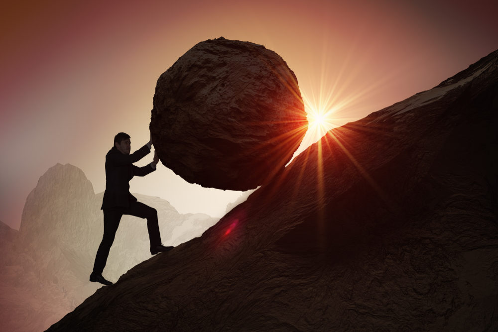 Silhouette of a businessman pushing a large boulder up a hill