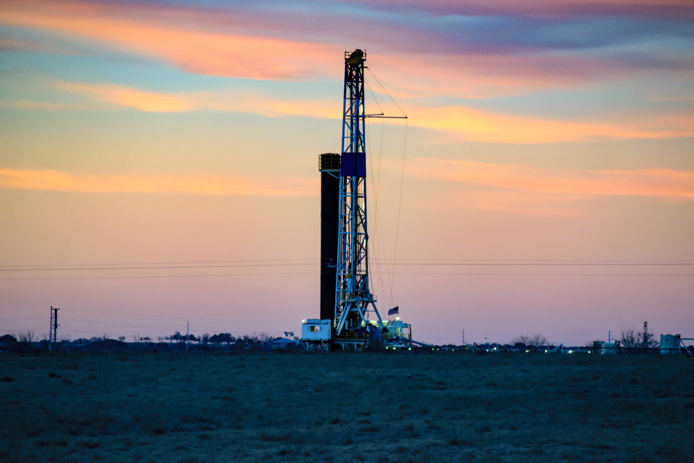 A tall fracking drilling rig stands against a sunset