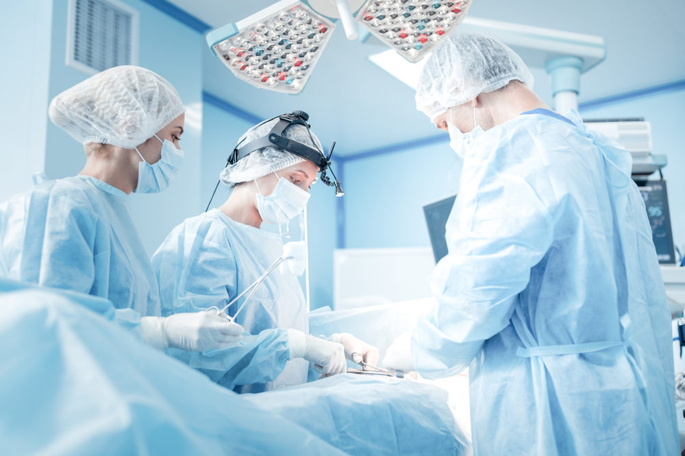 One male and two female doctors in gowns surround a patient on an operating table