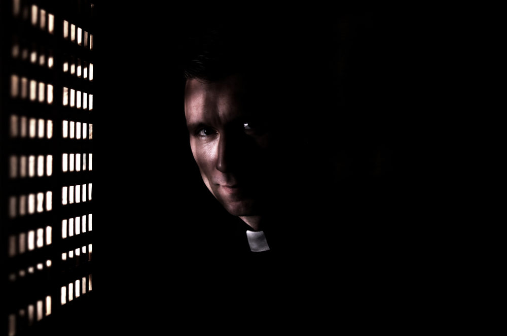 A priest in a confessional booth, looking at the camera