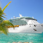 A large luxury cruise ship sails past a palm tree in turquoise blue waters