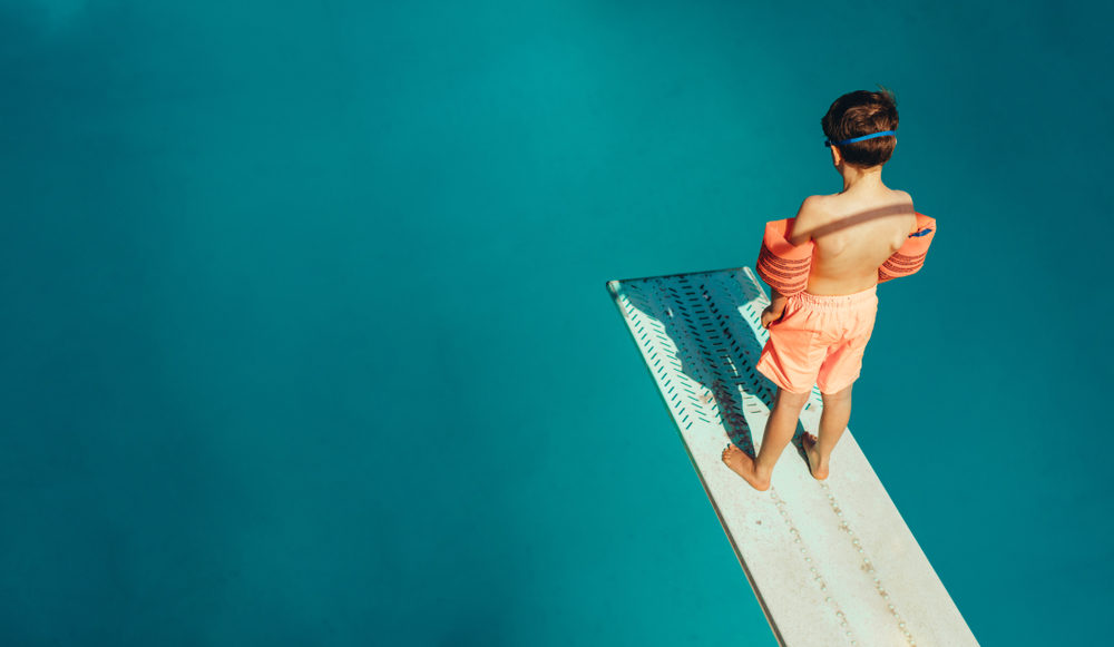 A young boy stands at the end of a springboard looking into the pool