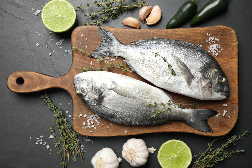 Two raw dorada fish on a cutting board with herbs and spices