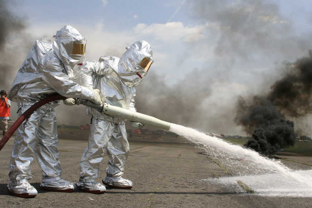 Two firefighters in protective suits spraying chemicals at an airport with smoke in the background