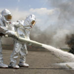 Two firefighters in protective suits spraying chemicals at an airport with smoke in the background