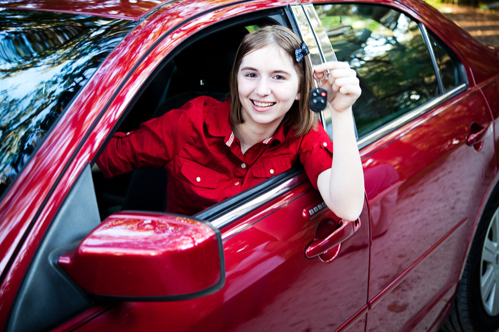 A young teenaged girl smiles in the driver's seat of a red car while holding a set of car keys