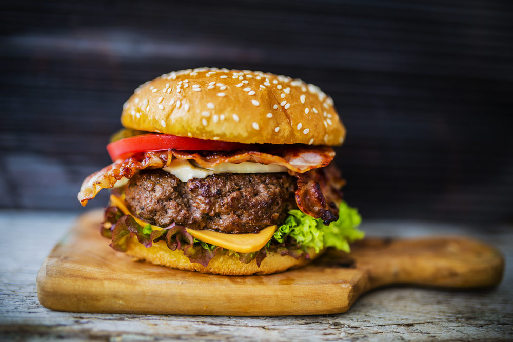 A large, greasy bacon cheeseburger on a wooden cutting board