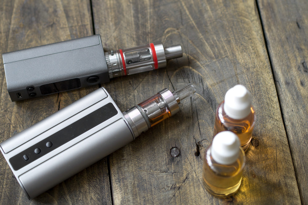 Two electronic vape pens on a table beside two vials of murky liquid