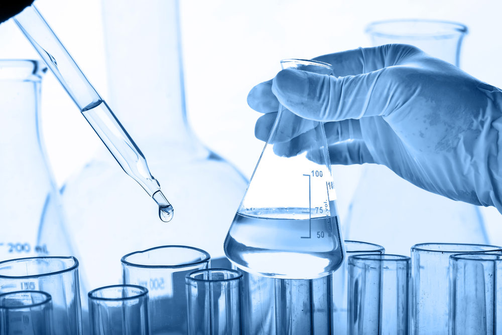 A gloved hand holds a flask of clear liquid with other vessels in the background