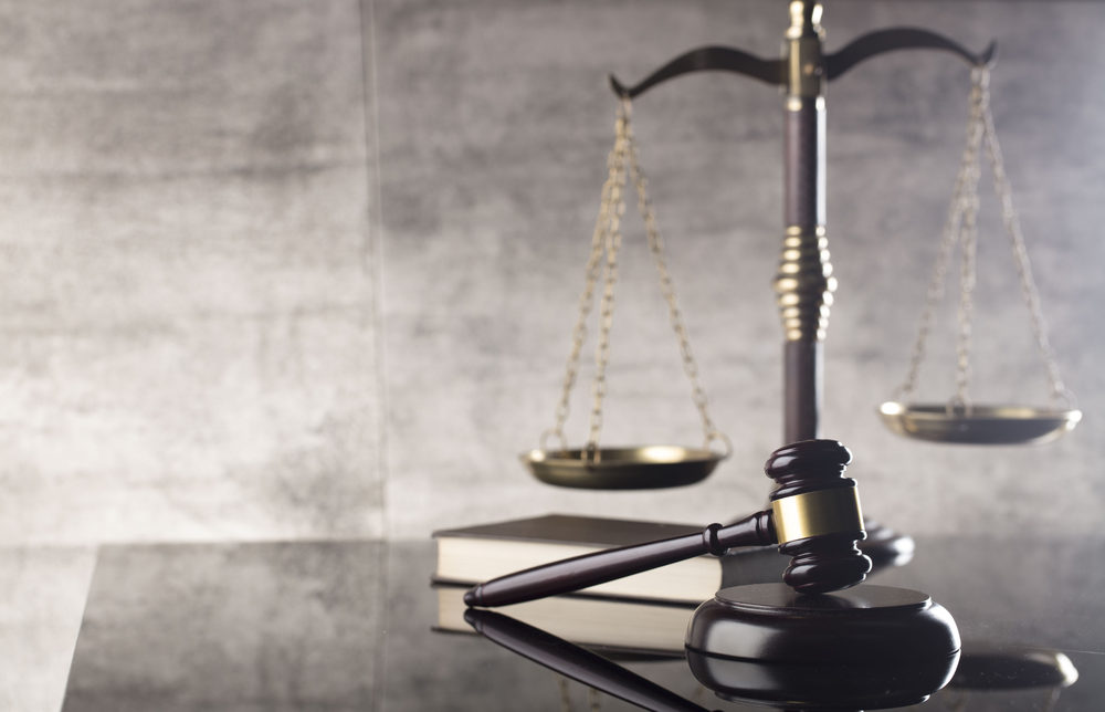 A dark brown gavel and block, with a book and the Scales of Justice in the background