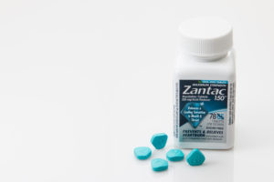 A bottle of Zantac 150 with some blue pills beside it, on a white background
