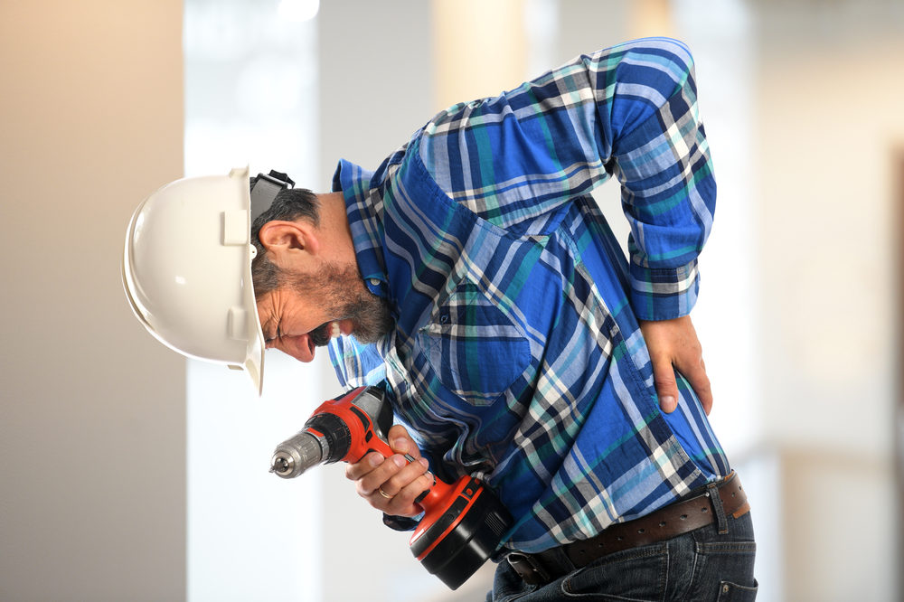 A construction worker holding a drill grimaces in pain and grabs his sore back