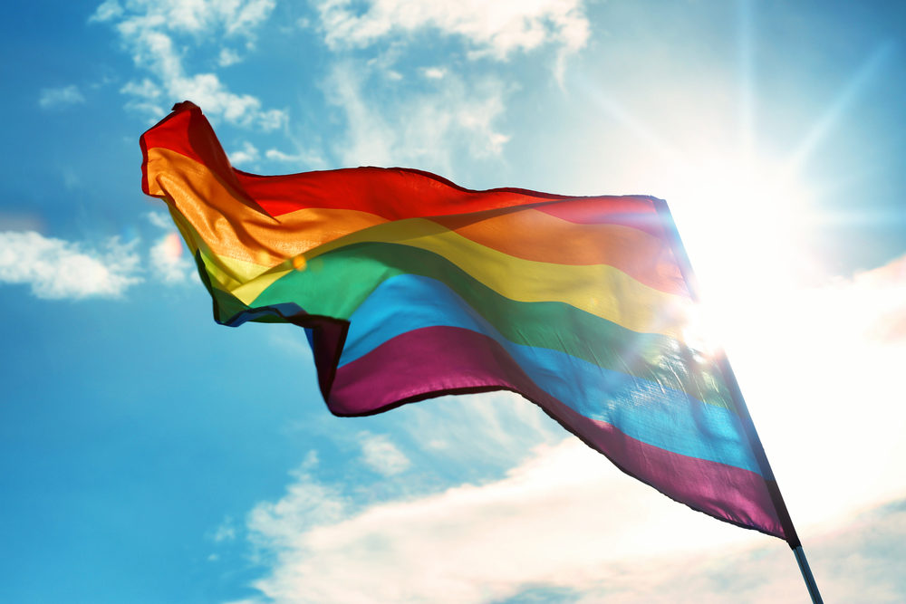 A rainbow flag flapping against a bright sun and blue sky