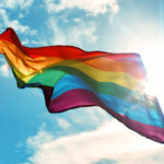 A rainbow flag flapping against a bright sun and blue sky