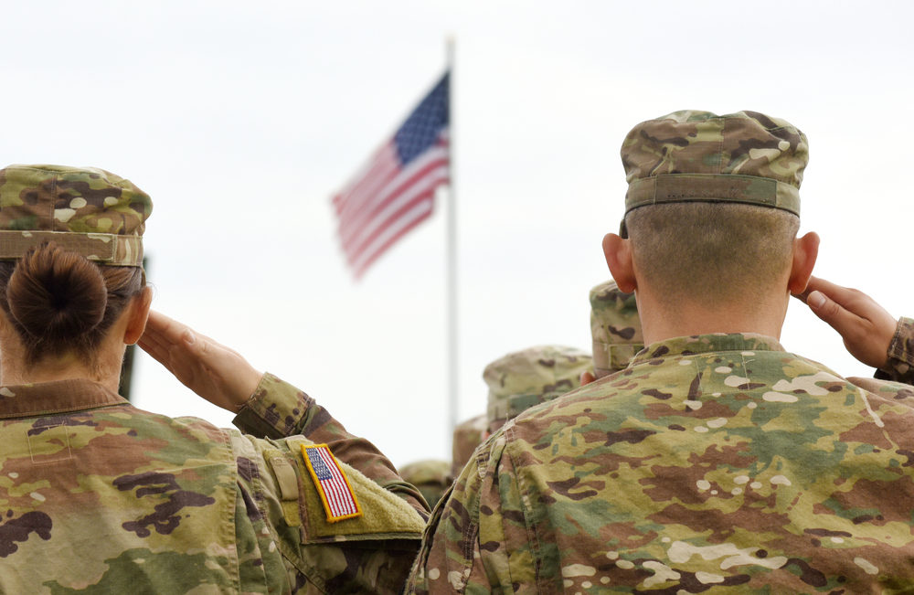 Male and female soldiers in uniform salute American flag outside.