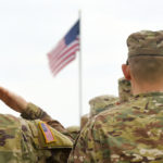 Male and female soldiers in uniform salute American flag outside.