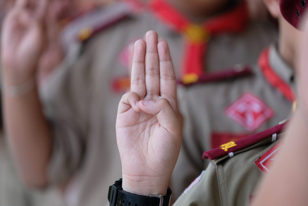 A boy in a Scout uniform holds up the traditional three-finger salute (the "Scout Sign")