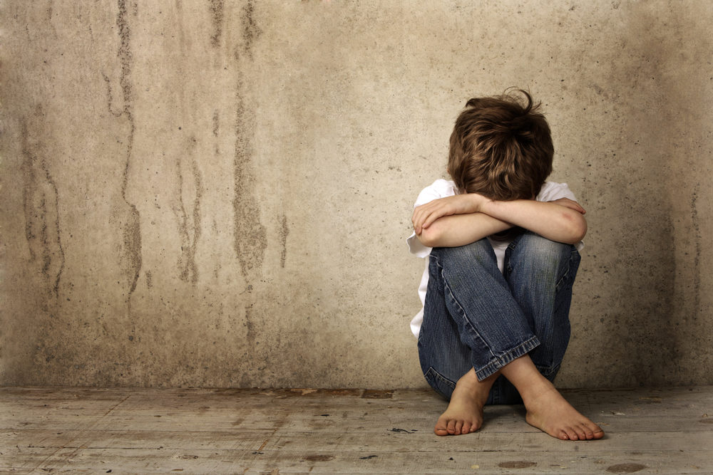 A young boy with his head in his hands sits alone and scared against a dirty wall