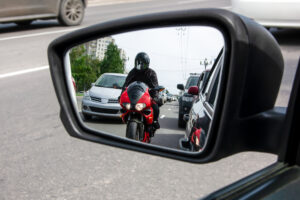 The side mirror of the car reflects a motorcyclist who is moving between cars
