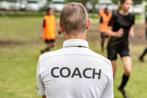 Back view of a male soccer, football, coach in white coach shirt watching his team play at an outdoor football field