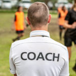 Back view of a male soccer, football, coach in white coach shirt watching his team play at an outdoor football field