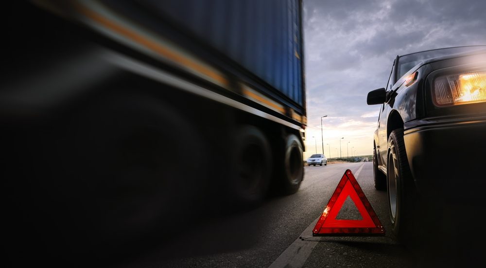 Red emergency stop sign (red triangle warning sign) and broken black SUV with a tractor trailer driving past on highway