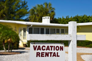 A Vacation Rental Sign in front of a yellow one story home on the beach.