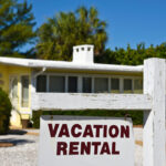 A Vacation Rental Sign in front of a yellow one story home on the beach.