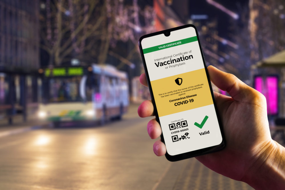 COVID-19 Vaccine Verification: Who Can and Can’t Require It? Smartphone displaying a valid digital vaccination certificate for COVID-19 in male's hand, downtown and city bus in background.