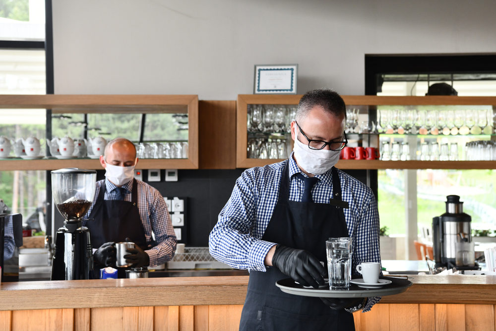 waiter in a medical protective mask serves the coffee in restaurant during coronavirus pandemic