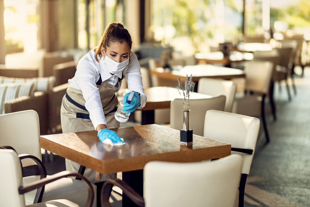 Young waitress disinfecting tables while wearing protective face mask ad gloves due to coronavirus epidemic.