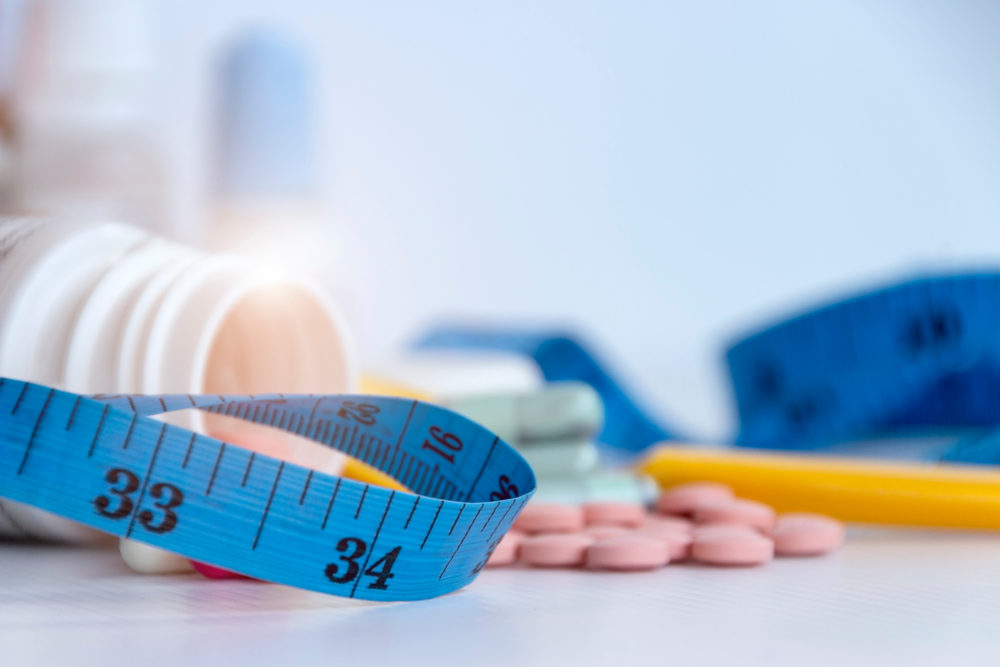 Weight loss pills spill from a bottle, with a blue measuring tape in the foreground