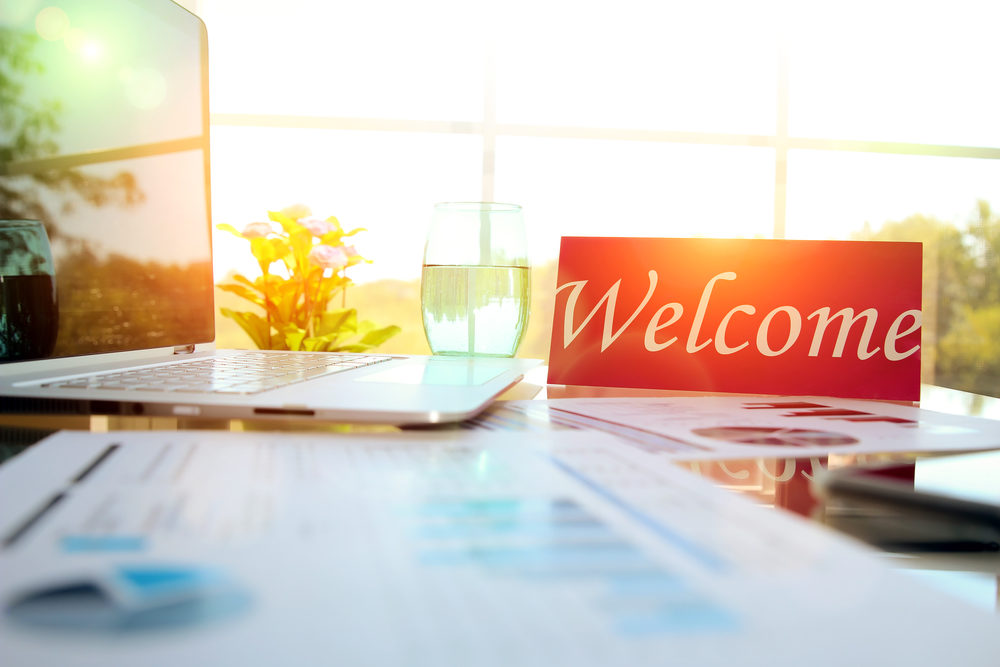 A "Welcome" sign sits on a cluttered office desk with a laptop and paperwork