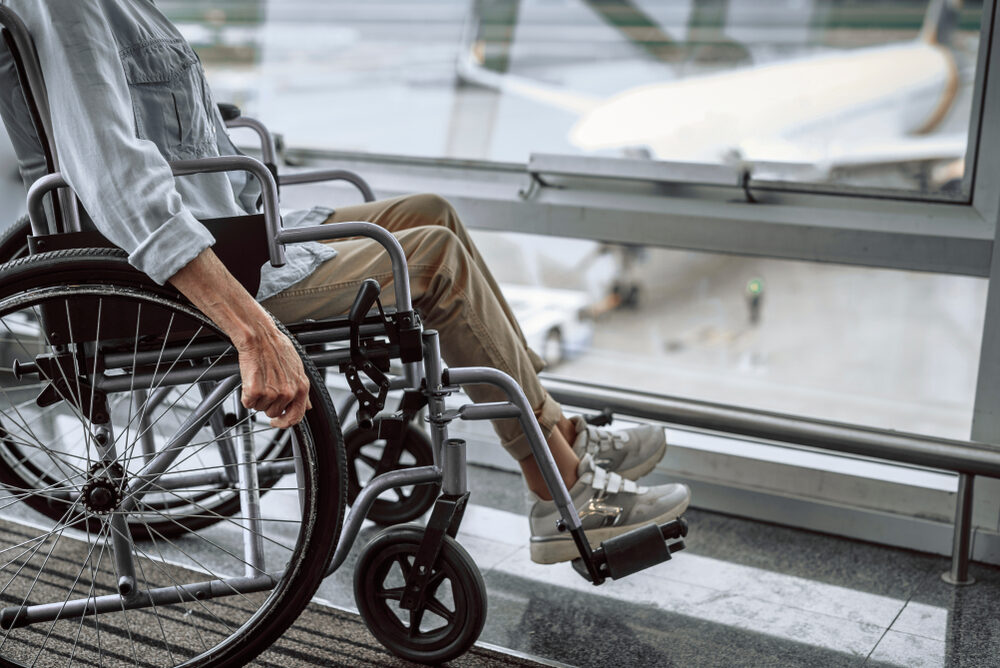 American Airlines Slapped with $50 Million Fine for Wheelchair Disability Violations Cropped photo of mature lady on wheelchair waiting in airport hall with airplane in the background
