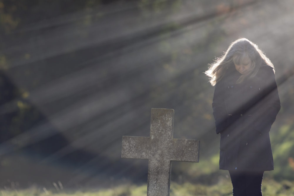 sunshine rays behind grieving widow in cemetery at stone cross grave site