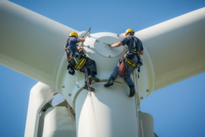 Inspection engineers preparing to rappel down a rotor blade of a wind turbine in a wind farm on a clear day with blue sky.