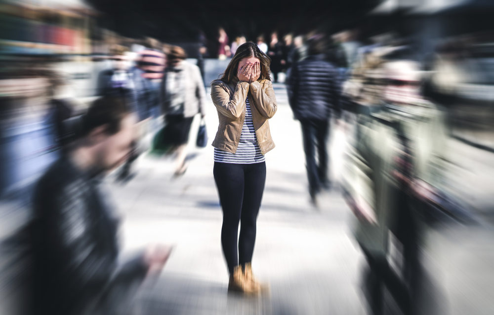 A woman holds her face in her hands as people move all around her (anxiety concept)