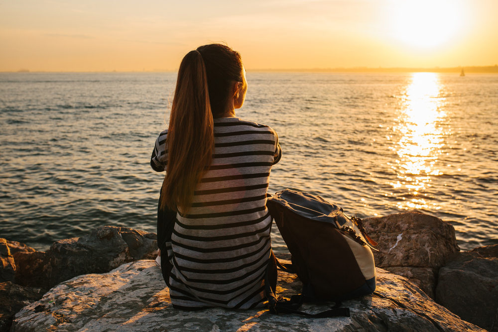 A young tourist girl with a backpack sits on the rocks next to the sea at sunset and looks into the distance. 