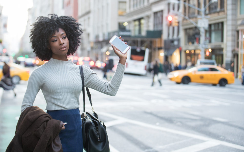 black woman holding her phone waiting for a rideshare to arrive in a city