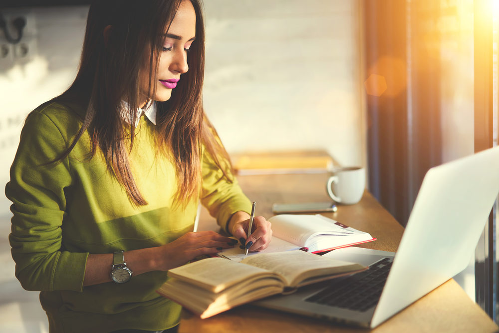 A young woman writes in a notebook while looking at an open book and laptop