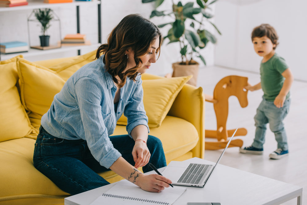 A woman works on a laptop in her living room while a child plays in the background