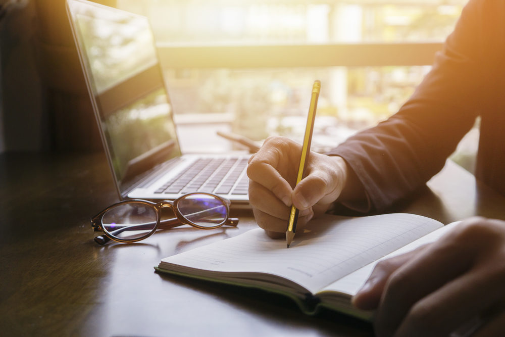 Close-up of a notebook being written in, with a laptop and reading glasses in the background