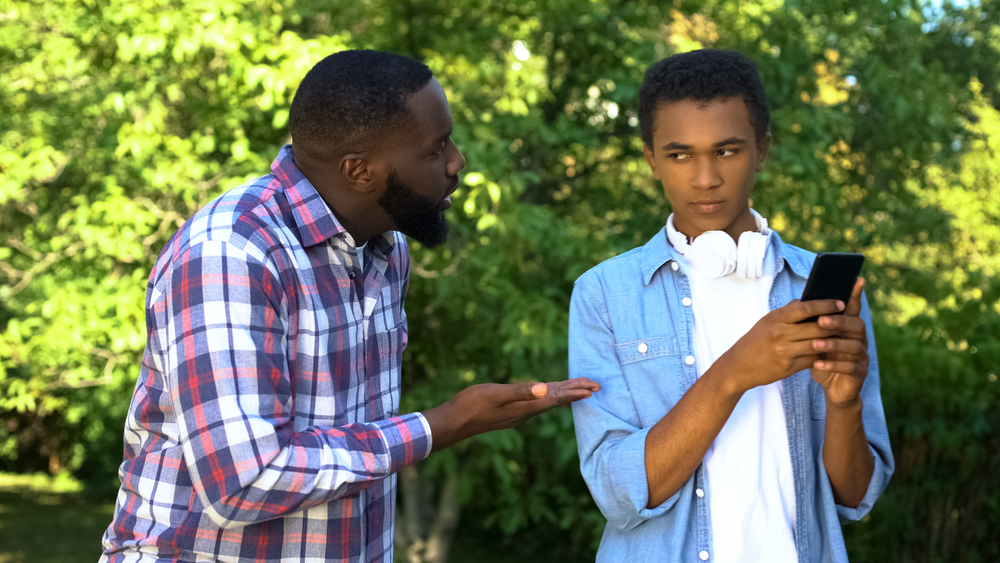 Father scolding male teen son playing smartphone game ignoring parent