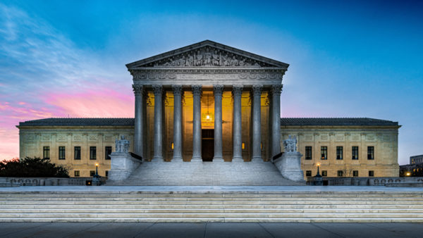 Ominous shadows on the front of illuminated United States Supreme Court building in Washington DC with sunrise clouds and sky in background and marble steps in foreground.