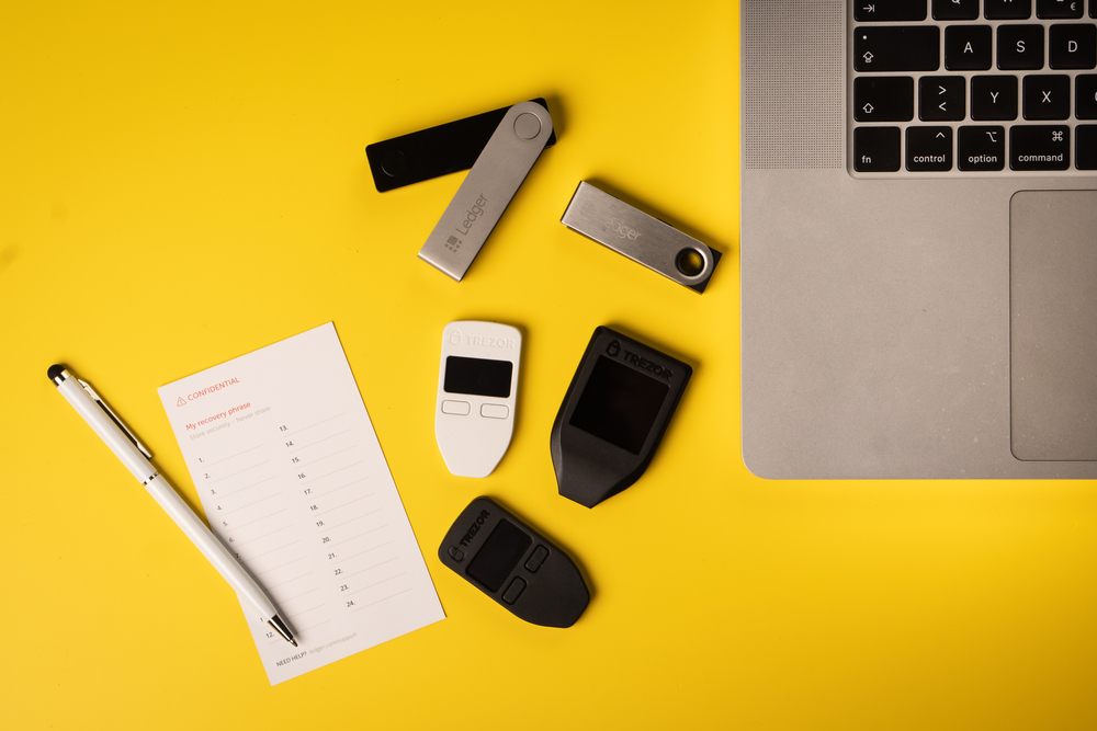 Top view of different cryptocurrency hardware wallets with paper recovery seed and laptop on the yellow background