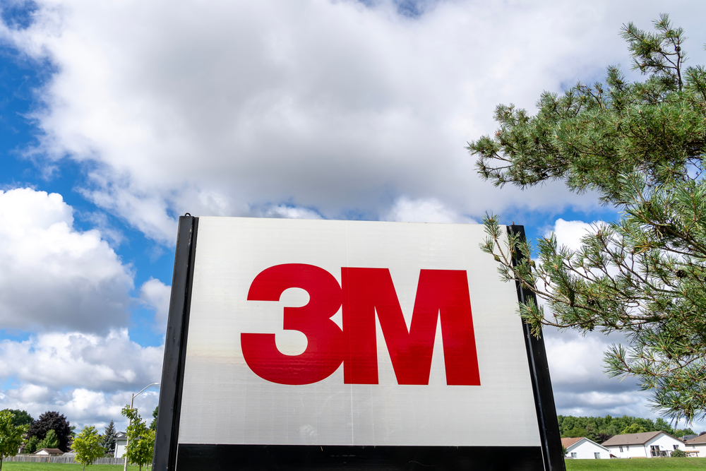 Close up of 3M sign with tree and blue sky in background.