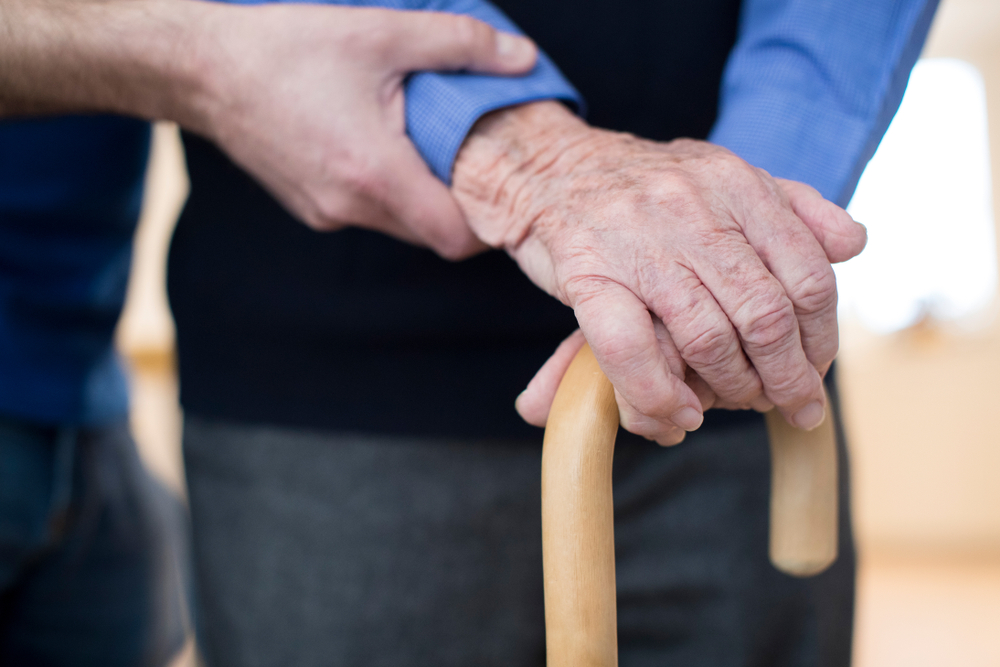 Senior Man's Hands On Walking Stick With Care Worker In Background