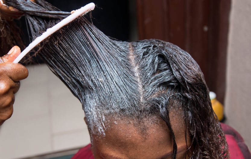 Hairdresser relaxing the hair on an African woman head and also using comb to stretch and apply the relaxer cream through the hair