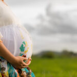 Pregnant woman wearing floral white dress affectionately holding her belly outside with newly planted rice field and cloudy sky in background.